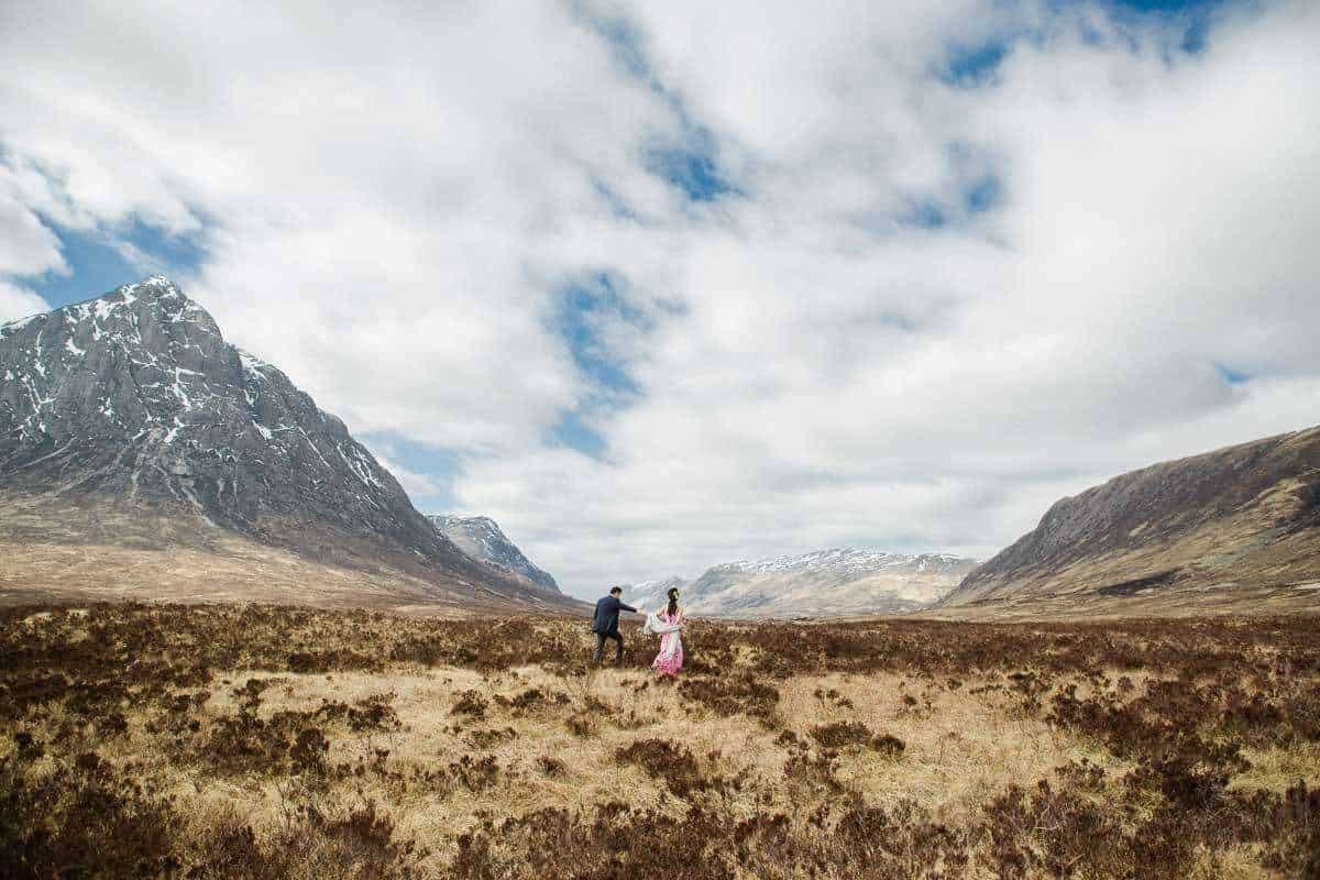A couple in formal attire walks hand in hand across a vast, open landscape with rugged mountains on both sides. The sky is covered with scattered clouds, and the terrain is a mix of grass and shrubs, creating a serene, natural setting.