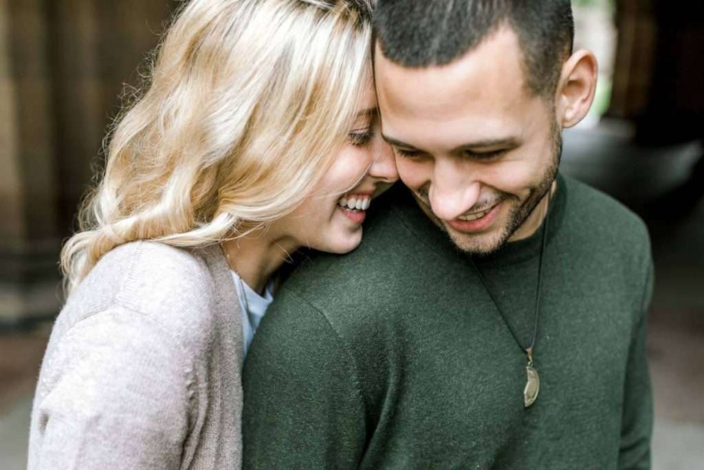 A smiling couple is embracing closely. The woman, with long blonde hair, is leaning on the mans shoulder, whispering something to him. The man, with short dark hair and a beard, is wearing a green sweater with a necklace. They look happy and relaxed.
