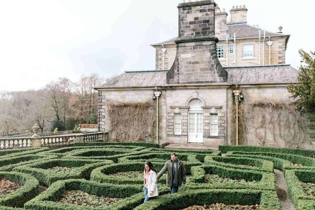 A couple walks through a green hedge maze in front of a historic stone building with a white door and small windows. The overcast sky creates a muted atmosphere, and autumn trees are visible in the background.