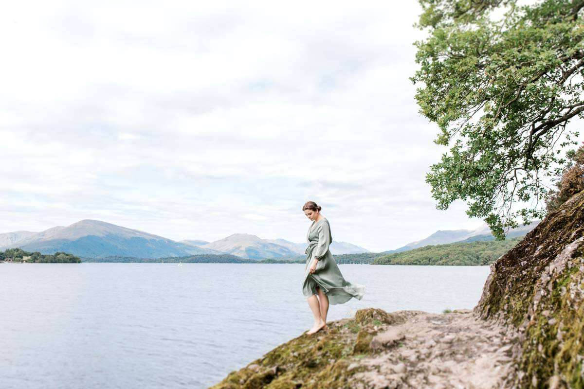 A woman in a flowing green dress stands on a rocky outcrop by a vast lake. Mountains and a cloudy sky form the background, while a tree extends into the frame from the right.