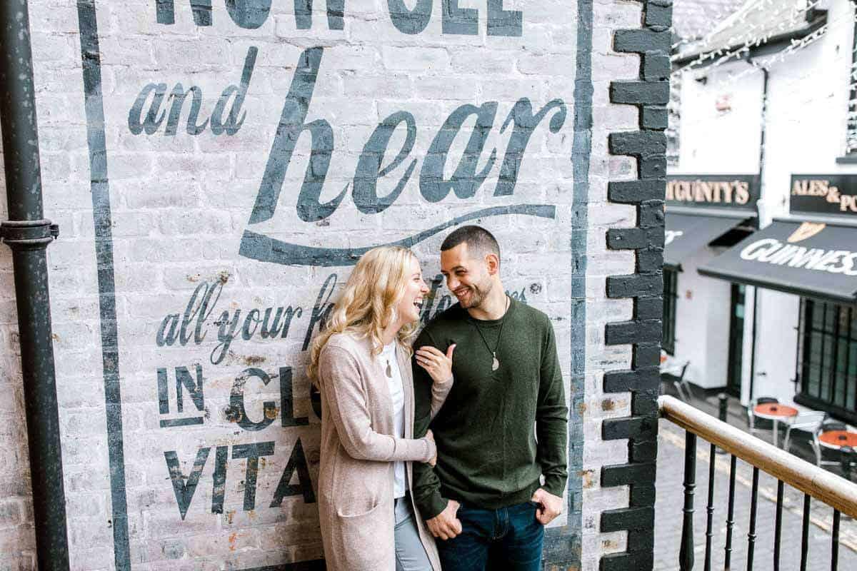 A couple stands smiling and leaning against a brick wall with vintage-style text. The background features a pub with Guinness signage, outdoor tables, and festive string lights. The man wears a green sweater, and the woman wears a light cardigan.