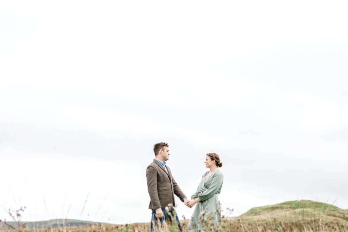 A couple stands holding hands in an open field under an overcast sky. The man is wearing a brown suit, and the woman is in a green dress. The landscape is minimal, with rolling hills in the background.