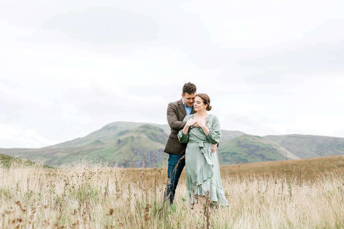 In the heart of Pollok Park, a couple stands affectionately in a grassy field with rolling hills beyond. The man dons a suit jacket, while the woman is elegantly clad in a light green dress—capturing the serene essence of their engagement shoot beneath an overcast sky.