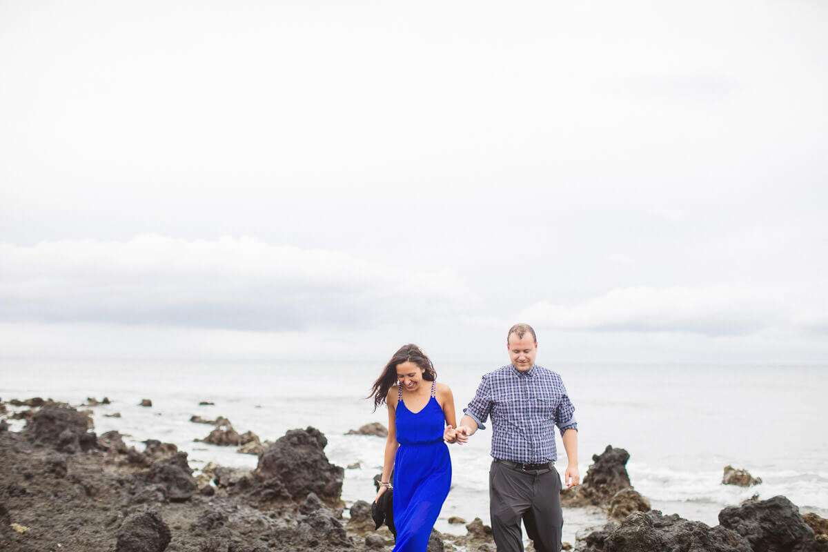 A couple is walking hand in hand along a rocky shoreline. The woman is wearing a bright blue dress, and the man is in a checked shirt and dark pants. The ocean and a cloudy sky are in the background.