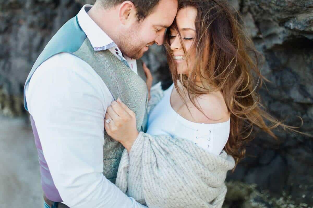 A couple stands closely together in an intimate embrace, smiling with their foreheads touching. The womans hair is windswept, and she wears a light shawl. They are near a rocky background, creating a romantic and natural setting.