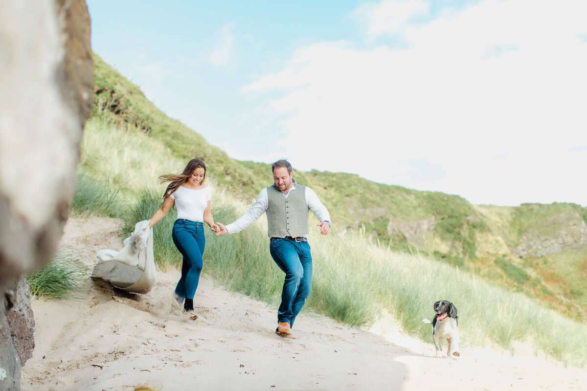 A couple holds hands and walks down a sandy hill, smiling and dressed in casual clothes. A dog runs beside them on the right. The background features green grass and a bright blue sky with scattered clouds.