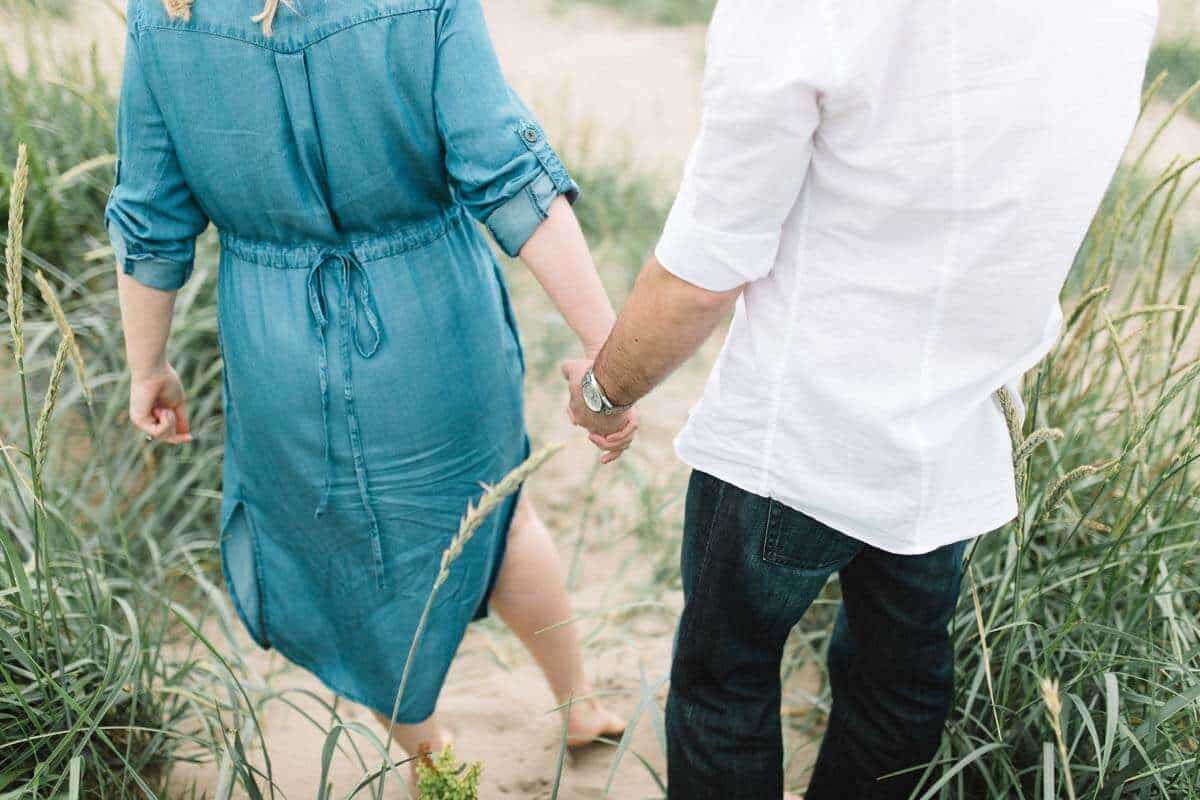 A couple holding hands walks through tall grass on a sandy path. The person on the left is wearing a blue denim dress, and the person on the right is wearing a white shirt and dark jeans. The scene conveys a calm, summery atmosphere.
