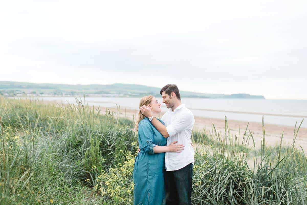 A couple stands close together, embracing and looking into each others eyes on a grassy beach. The background features a sandy shore and a calm, expansive sea under an overcast sky.
