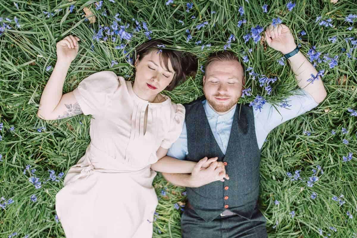 A couple lies on grass sprinkled with purple flowers, holding hands. The woman is wearing a light dress, and the man is in a vest and shirt. Both have their eyes closed, appearing peaceful and relaxed.