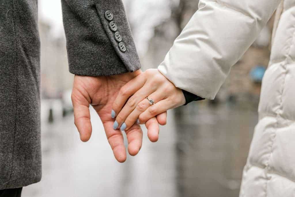 A couple holding hands outdoors. One person is wearing a gray coat, the other a white coat with a visible ring on their finger. The background is blurred, suggesting a city street on a rainy day.