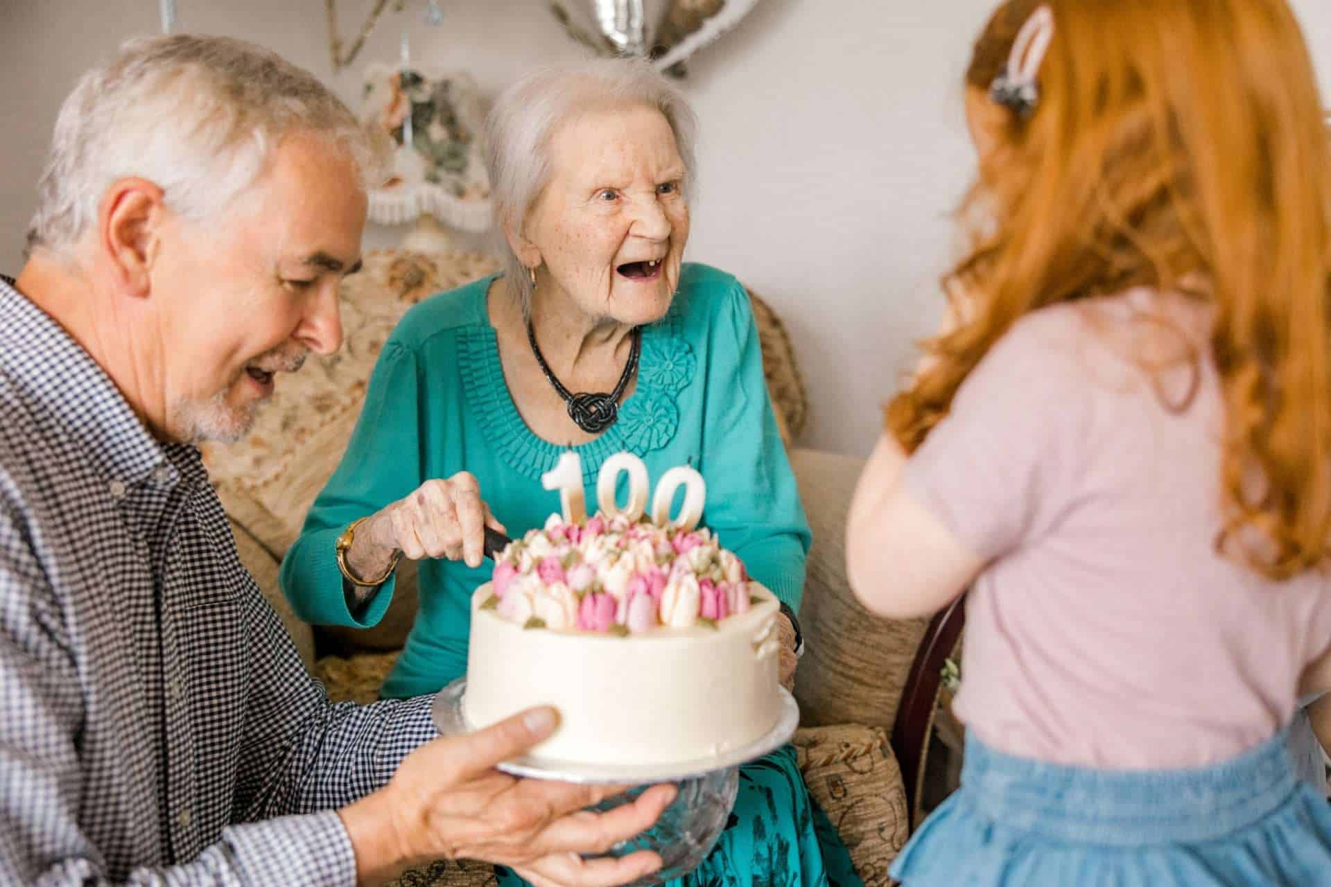 An elderly woman with a joyful expression sits in a chair, holding a cake with 100 candles. A man presents the cake to her, and a young girl with red hair stands nearby. The room has warm, celebratory decor.