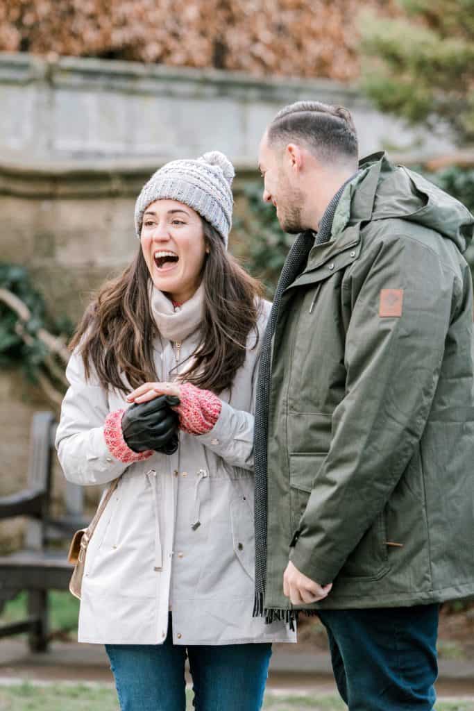 A woman and a man are outdoors, dressed in winter clothing. The woman, wearing a knit hat and gloves, is laughing, while the man, in a green jacket, looks at her smiling. The background is a garden setting with greenery and a wooden bench.