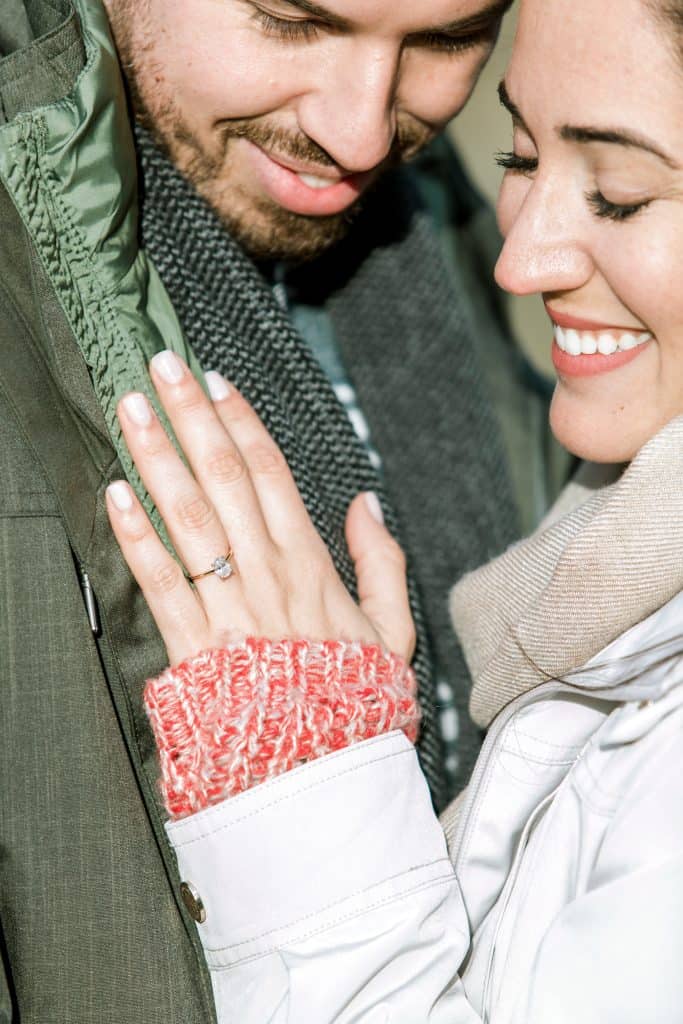 A close-up of a smiling couple. The woman is wearing a white coat and pink mittens, showing off an engagement ring on her hand. The man, dressed in a green jacket and gray scarf, is leaning in close, both looking content.