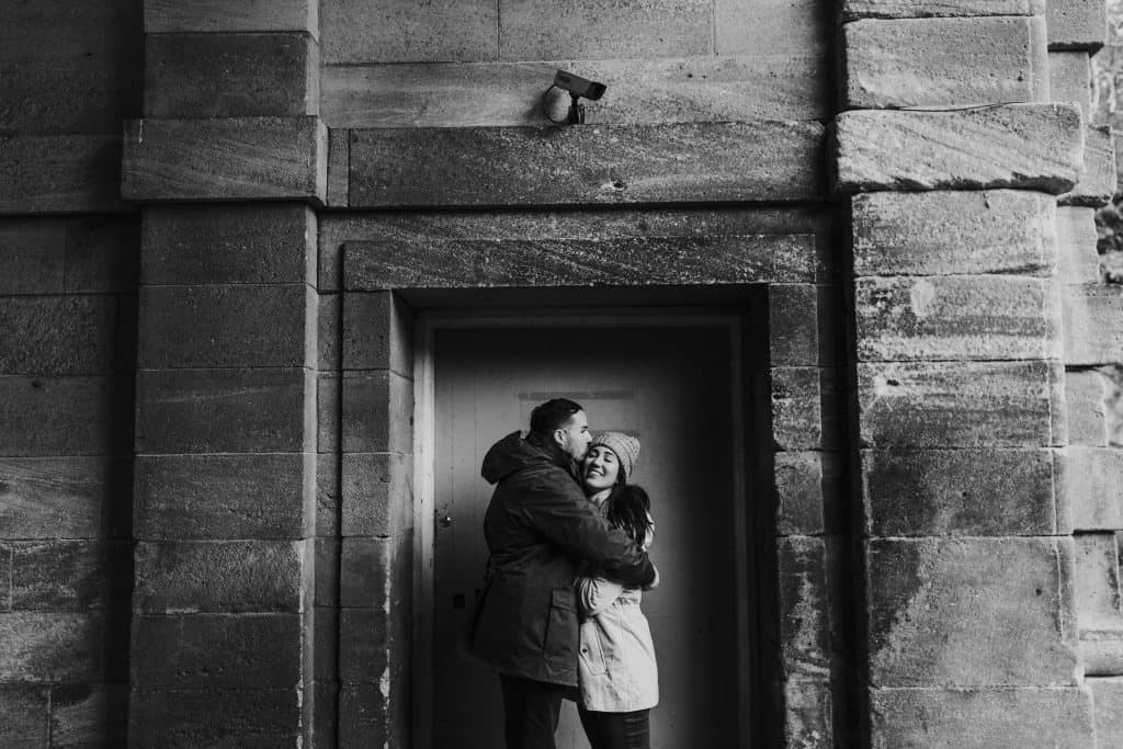 A couple embraces joyfully in a doorway framed by large stone blocks. The man wears a dark jacket, and the woman wears a light coat and knit hat. The setting has a rustic, historic feel with textured stone walls. Black and white photo.