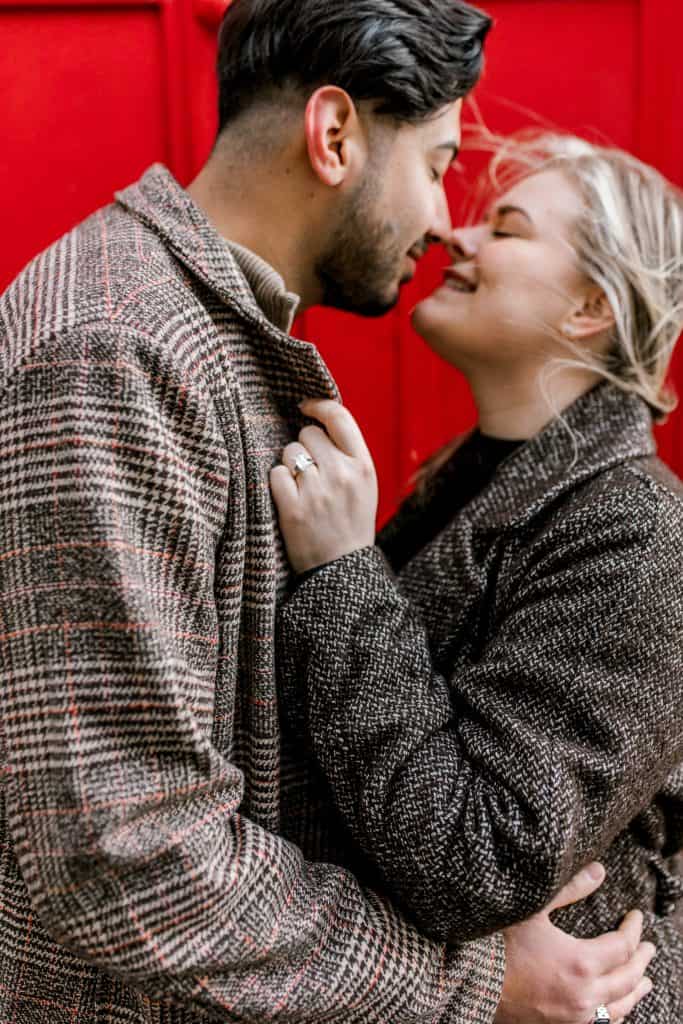 A couple wearing tweed coats stands close to each other, smiling with closed eyes. The woman is showing an engagement ring on her hand resting on the mans chest. A vibrant red background completes the scene.