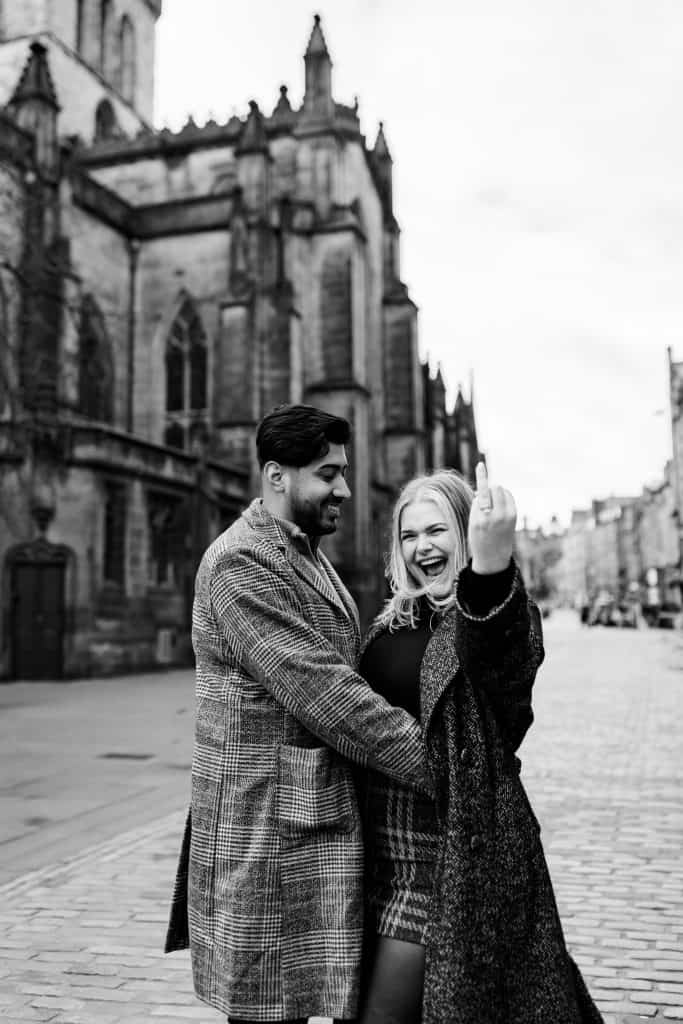 A joyful couple stands on a cobblestone street in front of an old, Gothic-style building. The woman smiles and shows off a ring on her finger. Both are dressed in winter coats. The scene is in black and white.