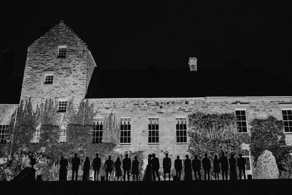 Silhouettes of a group of people, including a couple in the center, stand in front of a large stone building with illuminated windows at night. The building is covered with foliage, and the scene is captured in black and white.
