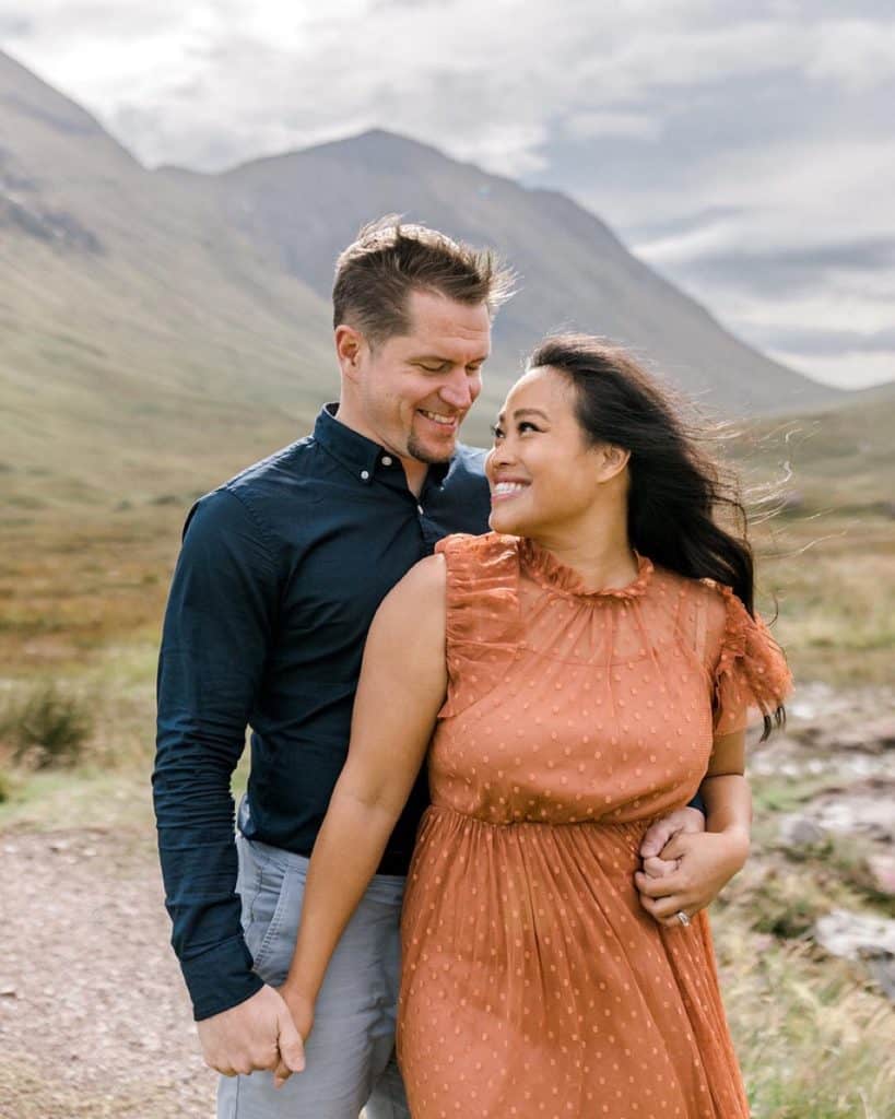 A couple stands on a dirt path in a scenic valley, holding hands. The woman wears a rust-colored dress, and the man wears a navy shirt and gray pants. Both are smiling, with mountains and a cloudy sky in the background.