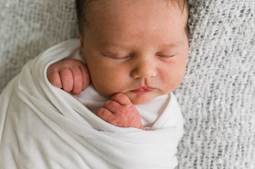 Newborn baby wrapped in a white blanket, peacefully sleeping on a textured gray surface. The babys hands are tucked near the face, and the eyes are closed.