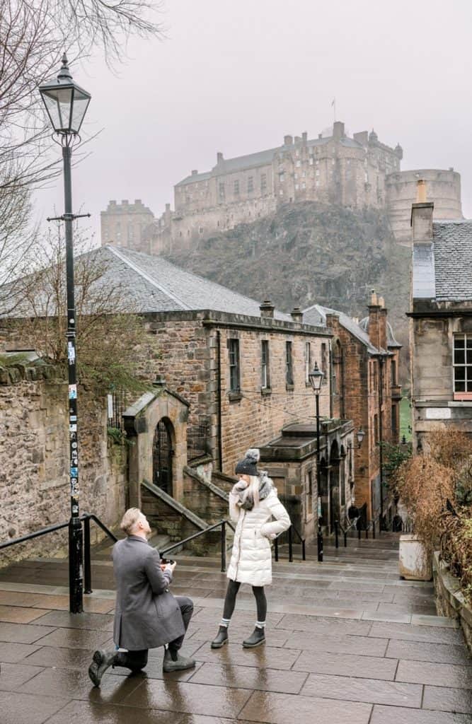 A person in a gray coat kneels on one knee, holding a ring box, proposing to another person in a white coat and hat. They are on a cobblestone street with a historic castle on a hill in the background on a foggy day.