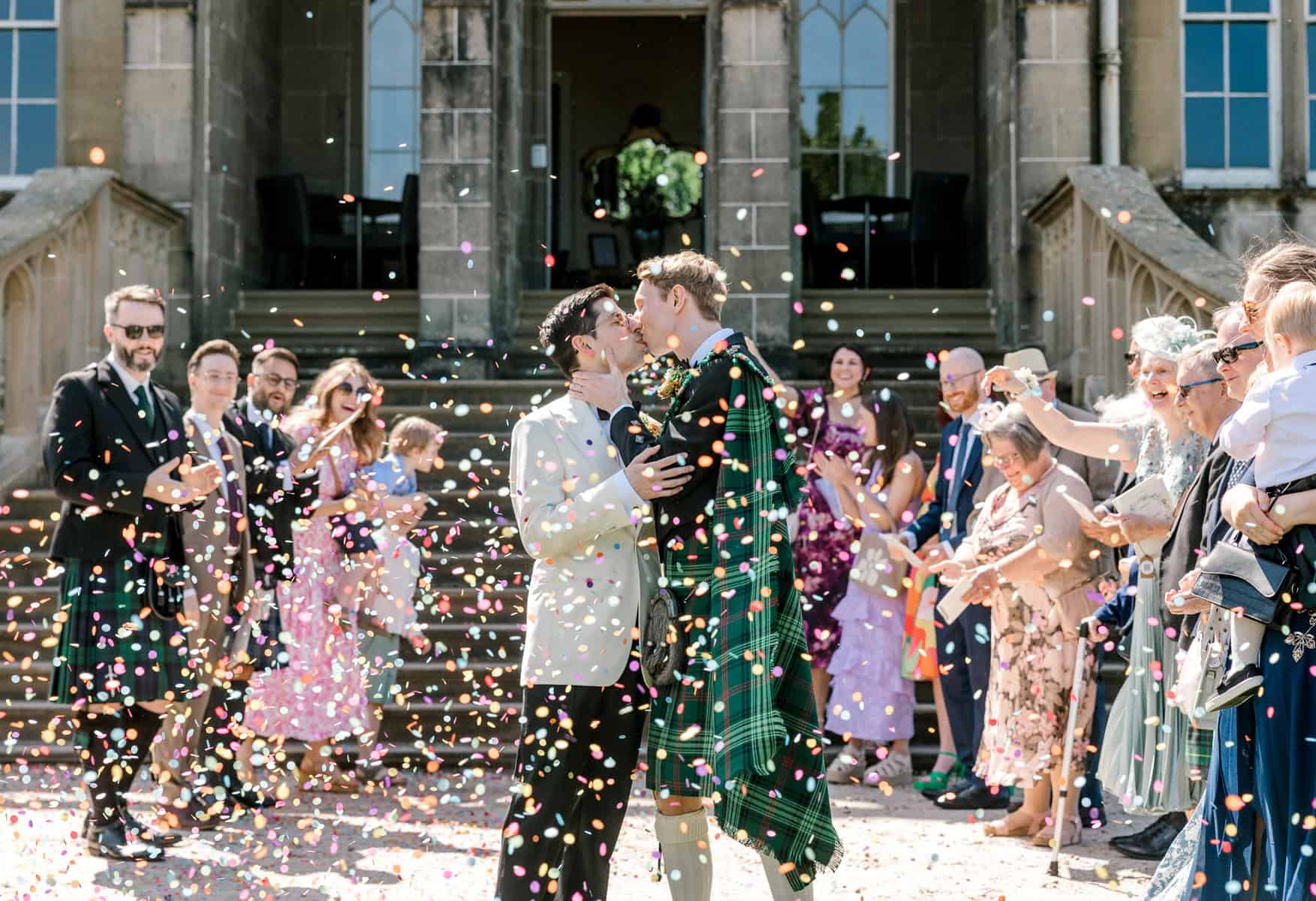Two grooms kiss outside a grand building as wedding guests in formal attire, some in kilts, celebrate and throw colorful confetti. People smile and clap, creating a joyful, festive atmosphere on the steps.