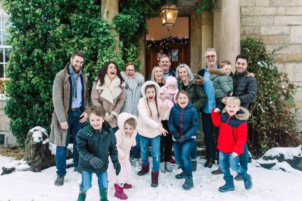 A group of adults and children dressed in winter clothes are smiling and posing together outside a house with snow on the ground. The house is decorated with greenery and fairy lights.