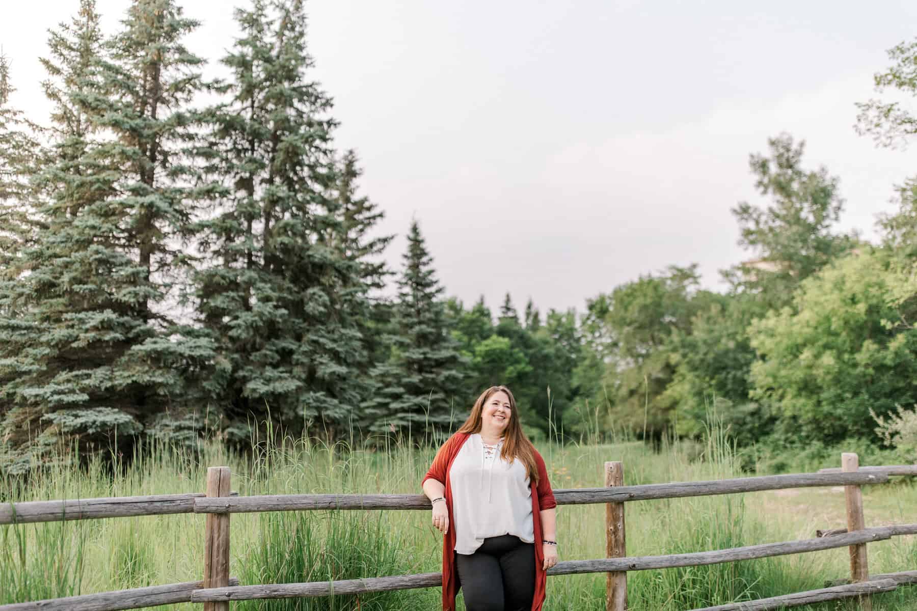 A woman with long brown hair, wearing a white top and red cardigan, stands smiling against a wooden fence. Behind her are tall green grass and pine trees under a clear sky.