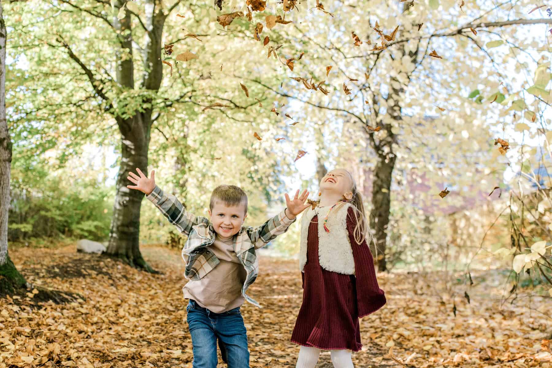 Two children play in an autumn forest, smiling and throwing fallen leaves into the air. The ground is covered with orange leaves, and sunlight filters through the trees, creating a joyful, playful scene.