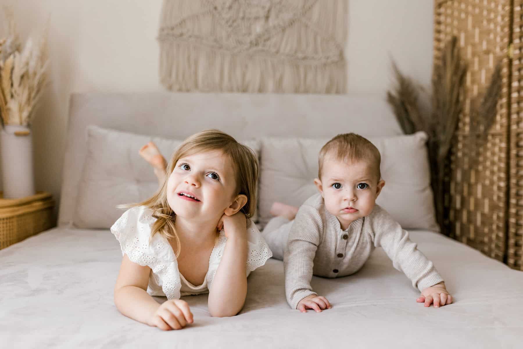 Two young children, a girl and a baby, lie on their stomachs on a bed. The girl is looking up and smiling while the baby looks forward with a neutral expression. The room has neutral decor and soft lighting.