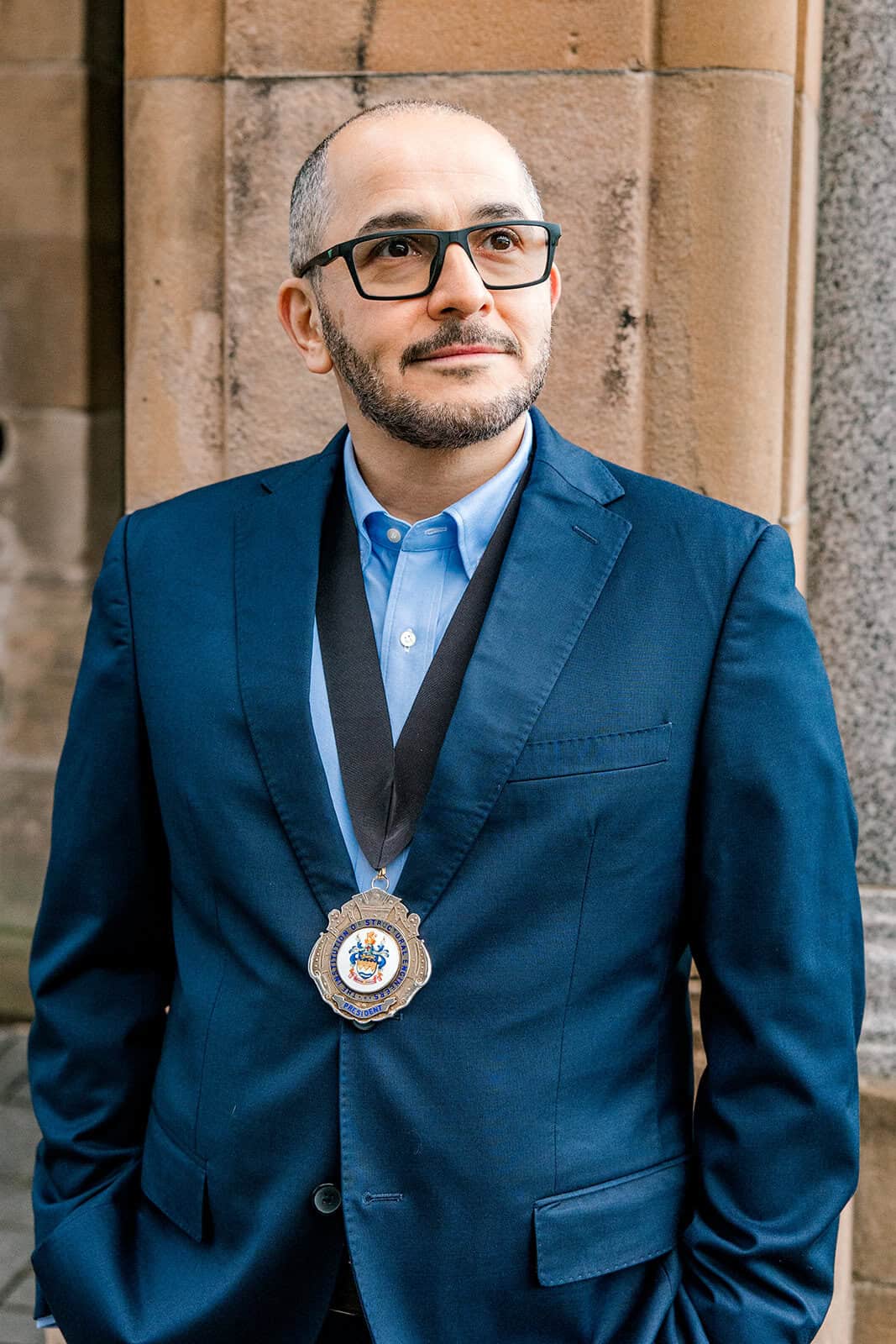 A man wearing glasses and a blue suit with a medallion around his neck stands in front of a stone building, looking slightly upward with a thoughtful expression.