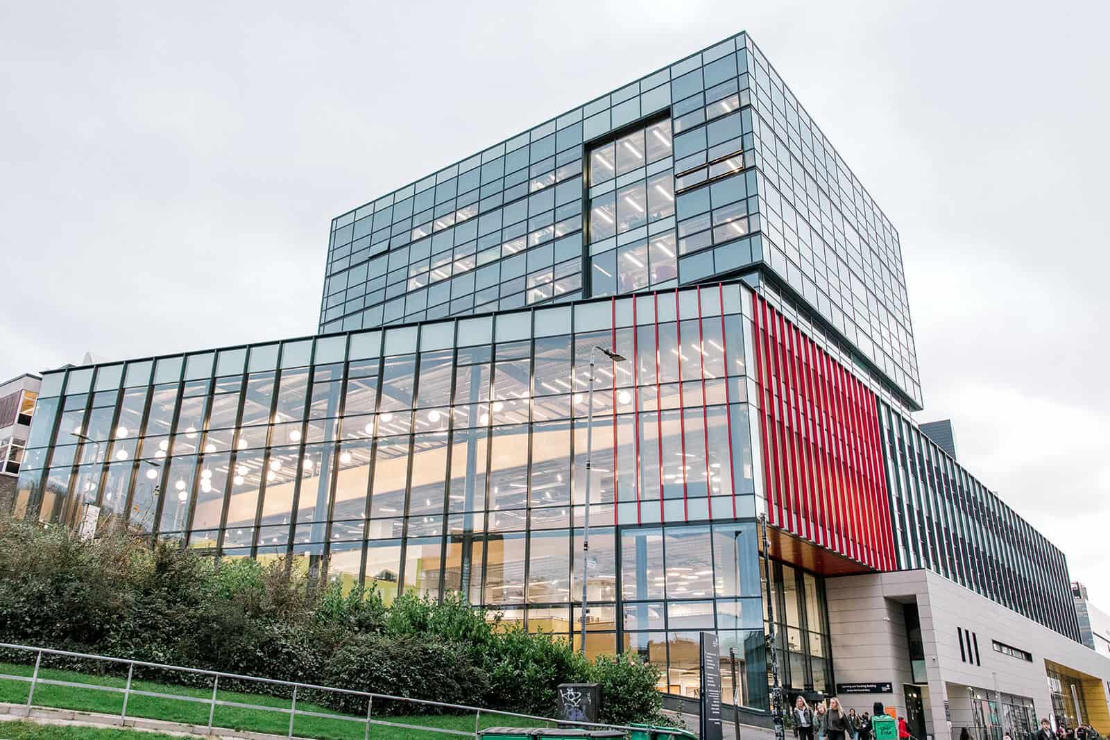A modern, multi-story glass building with a mix of transparent and red vertical panels. The lower floor is brightly lit, and greenery surrounds the entrance, with people walking outside under an overcast sky.