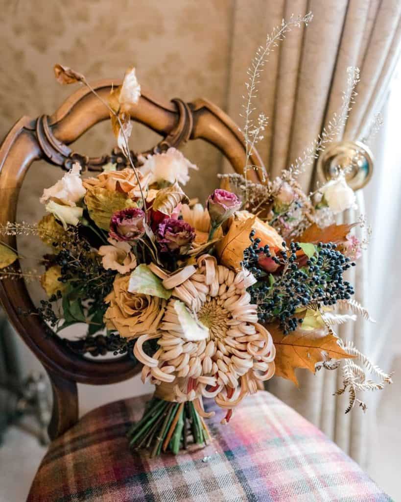 A bouquet of autumn flowers with dried leaves, berries, and various blooms rests on a vintage wooden chair with a checked cushioned seat, set against softly patterned wallpaper and pale curtains.