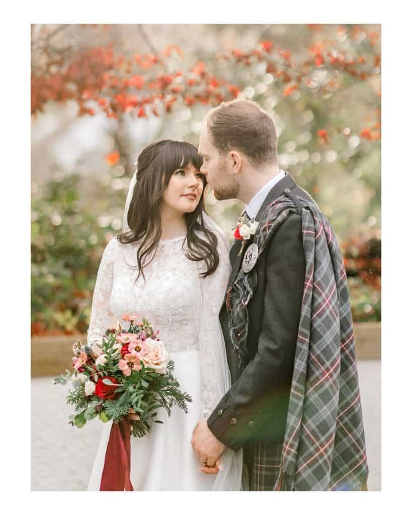 A bride in a white lace dress holds a bouquet whilst standing close to a groom in a kilt and tartan sash. They gaze lovingly at each other in an outdoor setting with red berries in the background.