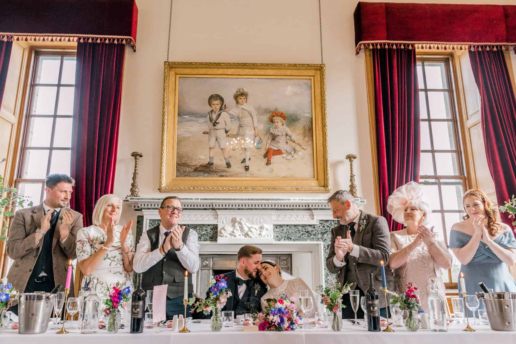 A wedding couple share a kiss at a banquet table surrounded by applauding guests. The group is seated in an elegant room with large red curtains, floral arrangements, and a painting of children above a fireplace in the background.