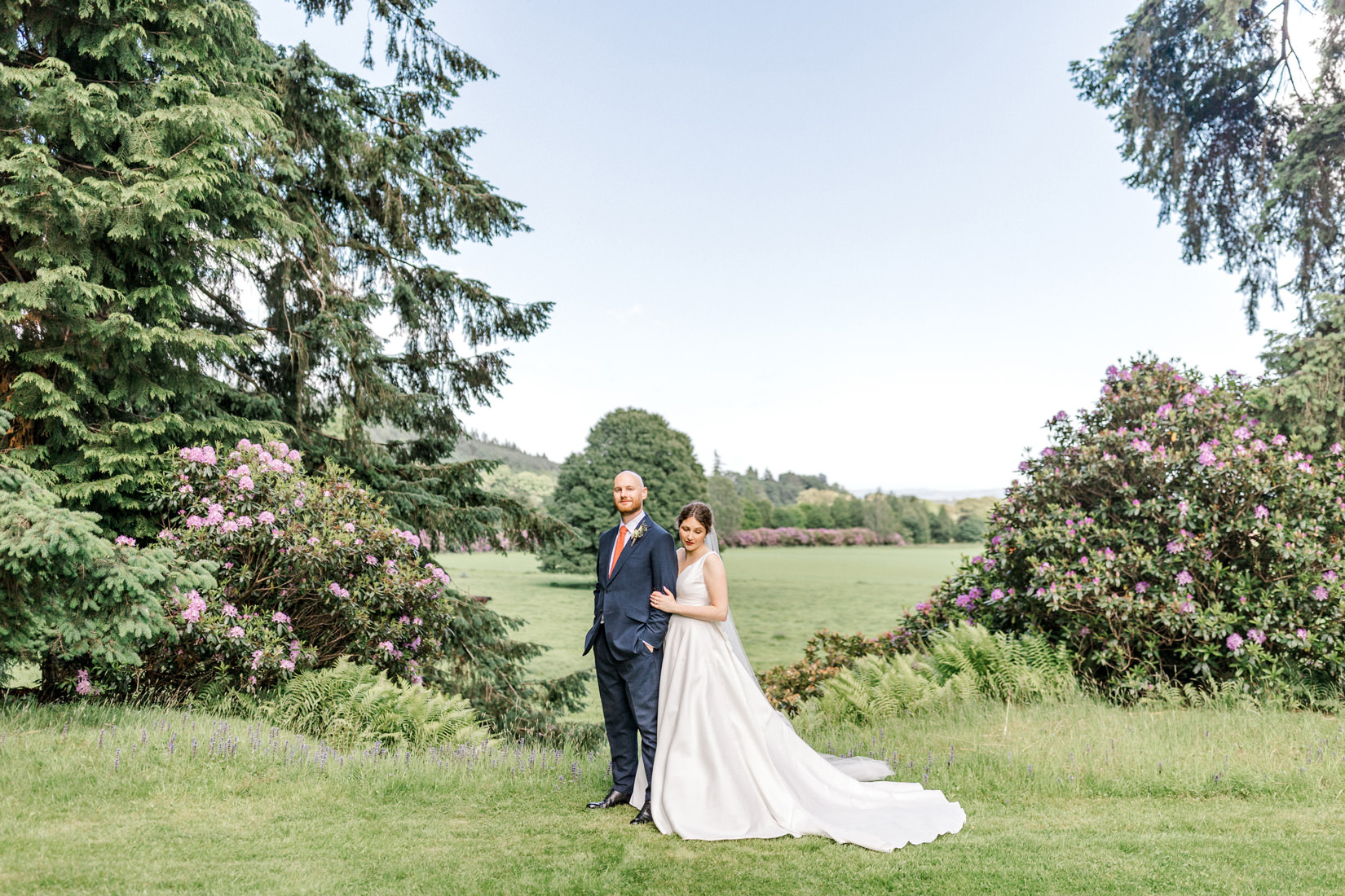 A bride in a white gown and a groom in a navy suit stand together on a lush green lawn surrounded by tall trees and flowering bushes under a clear sky.