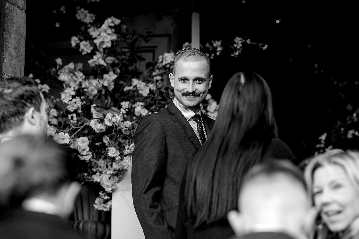 A man in a suit with a moustache smiles whilst standing among people at a formal event, with flowers in the background. The image is in black and white.