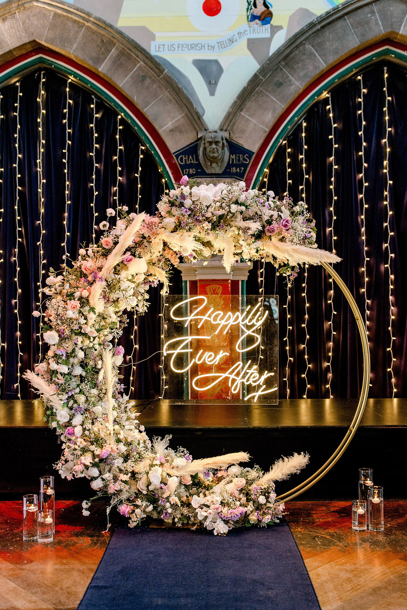 A circular floral arch decorated with pastel flowers and greenery, featuring a neon sign that reads “Happily Ever After.” The arch stands in front of dark curtains with fairy lights and glass candle holders on the floor.
