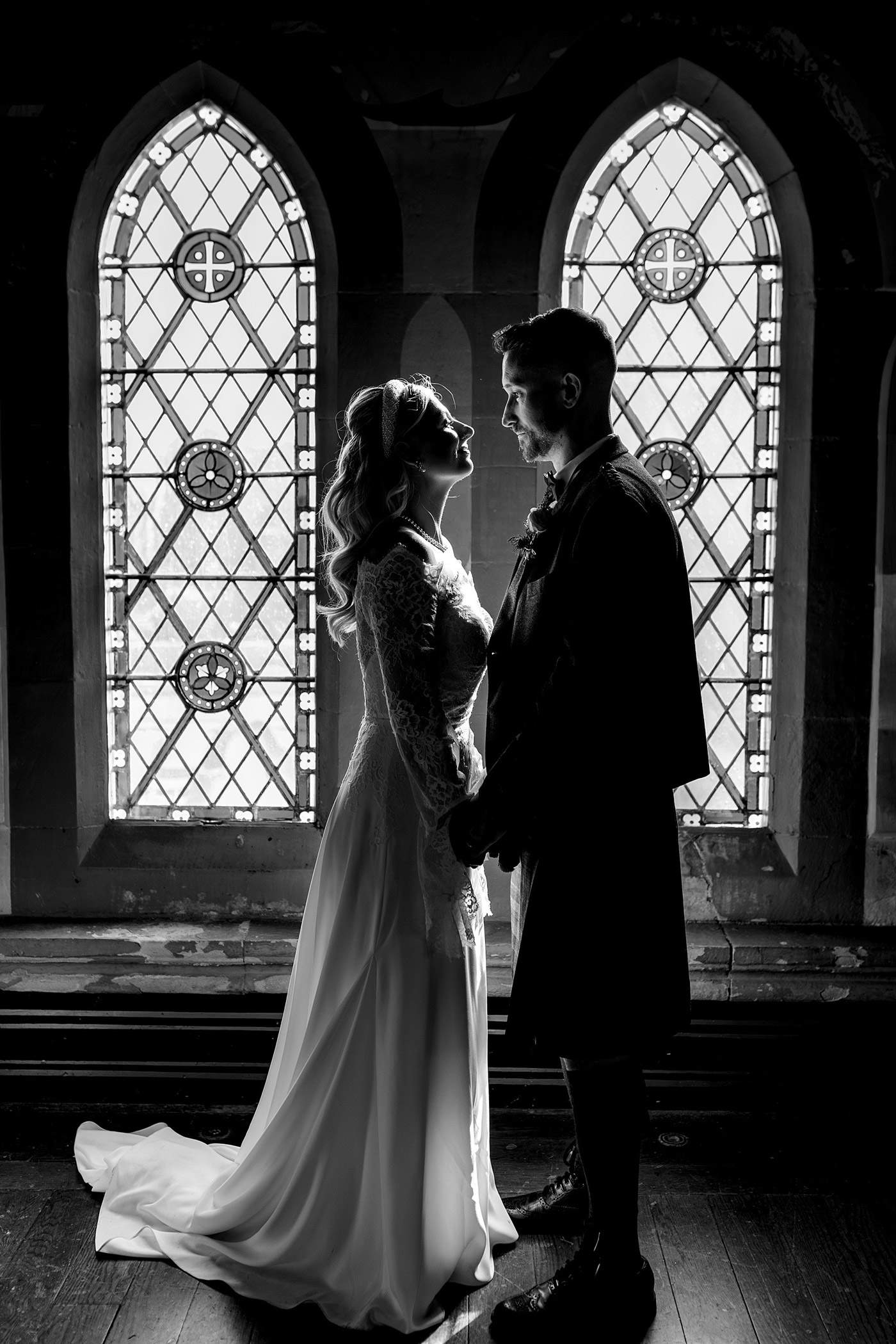 A bride and groom stand facing each other, holding hands in front of two tall, ornate stained-glass windows. The image is in black and white, giving a romantic, timeless feel.