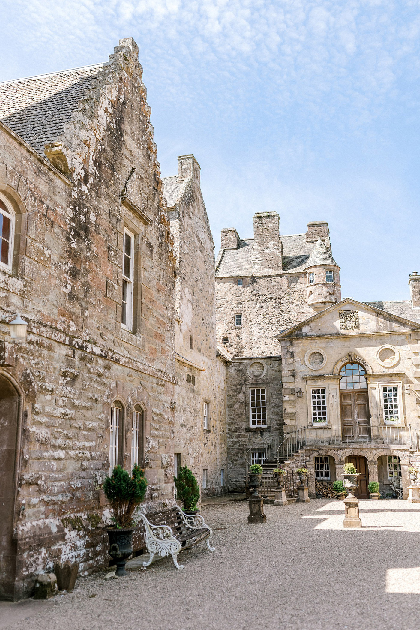 A courtyard of an old stone castle with arched windows, potted plants, ornate benches, and a staircase leading to an upper floor under a partly cloudy blue sky.