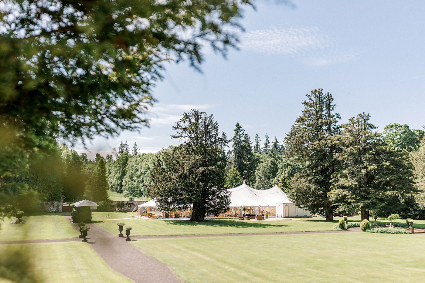 A large white event marquee is set up on a neatly mown lawn surrounded by tall trees and greenery under a clear blue sky. Paths and manicured hedges are visible in the foreground.