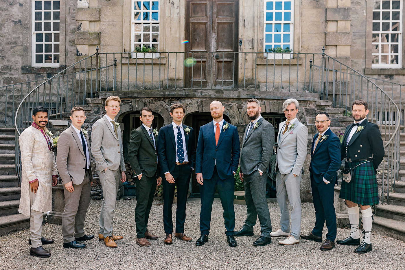 A group of ten men dressed in formal and semi-formal attire pose for a photo outside a stone building with large windows and a staircase.