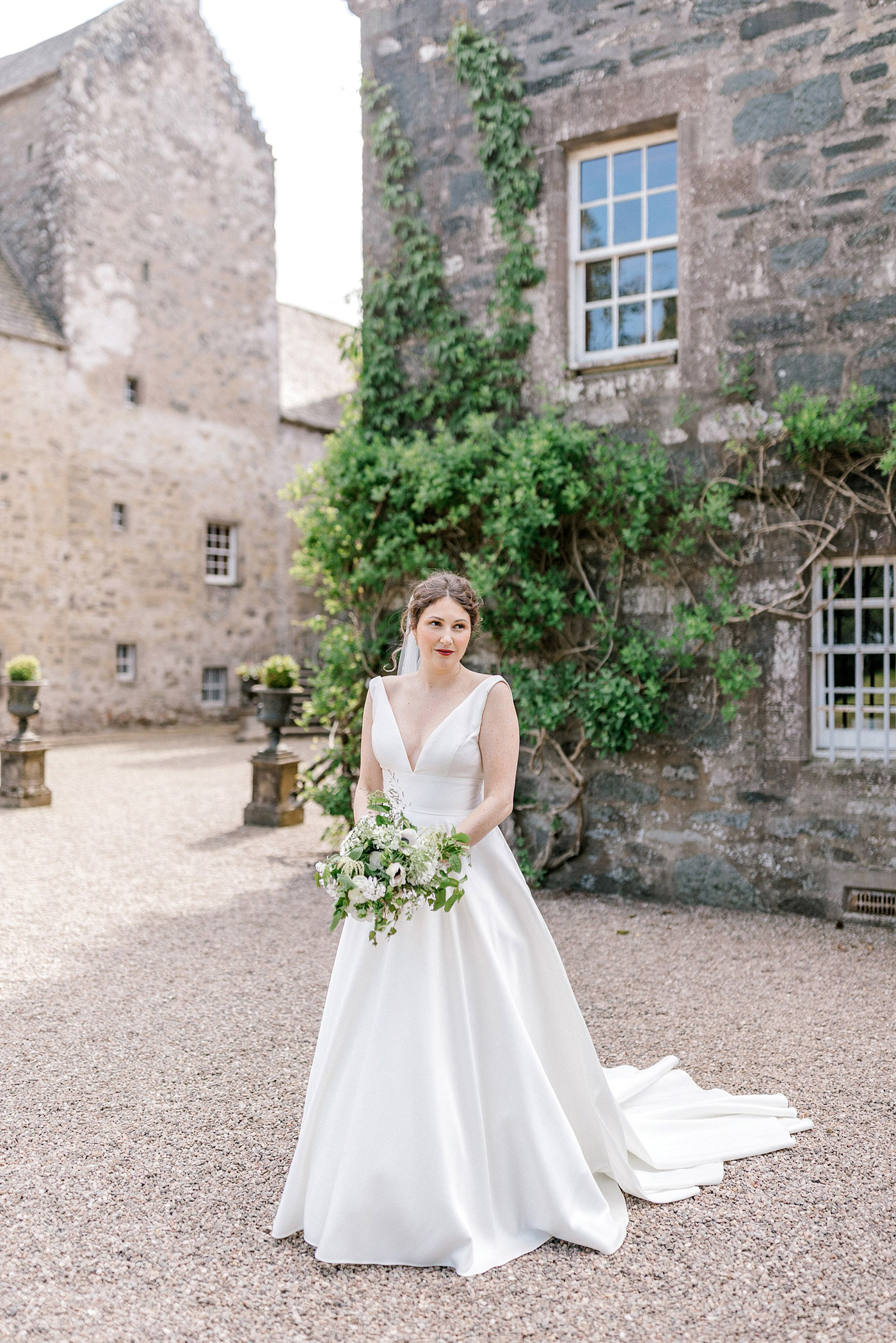 A bride in a white dress holding a bouquet of white and green flowers stands outside a stone building covered with ivy, on a sunny day.