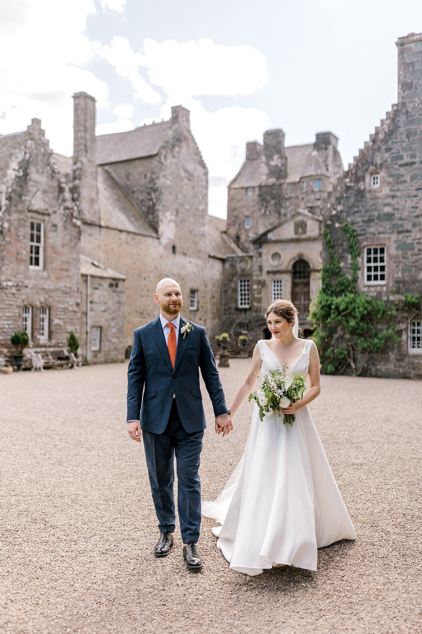 A bride in a white dress and a groom in a blue suit walk hand in hand outside a historic stone building on a bright day, with greenery and overcast sky in the background.