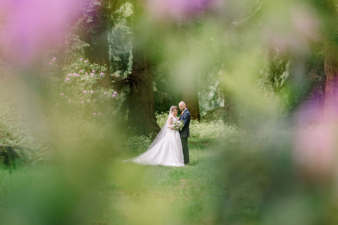 A bride and groom stand closely together in a lush, green forest, surrounded by soft-focus flowers and trees, creating a dreamy, romantic atmosphere.
