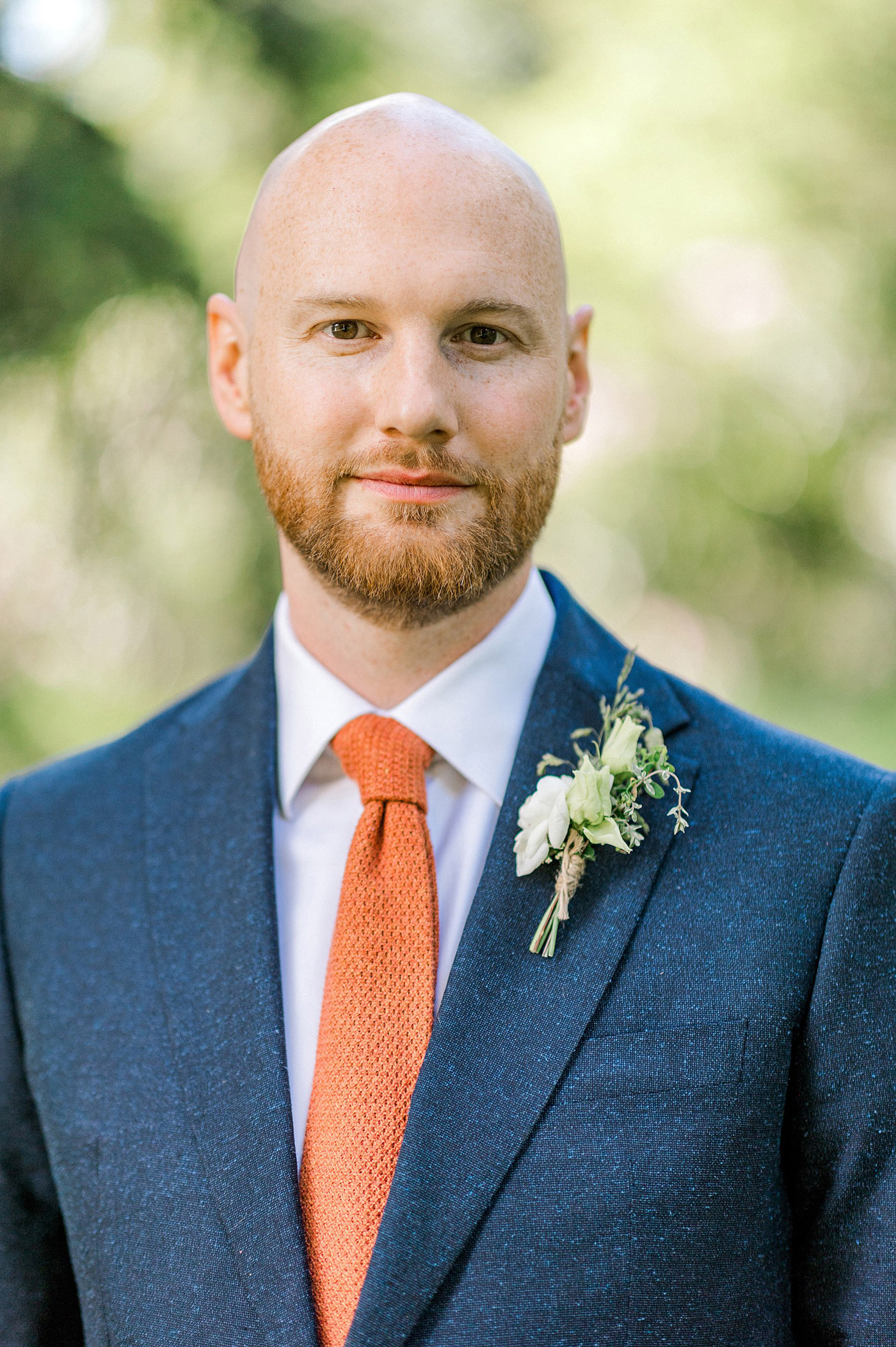 A man with a closely trimmed beard and bald head wears a blue suit, white shirt, and orange tie. He has a floral buttonhole pinned to his lapel and stands outdoors with greenery blurred in the background.