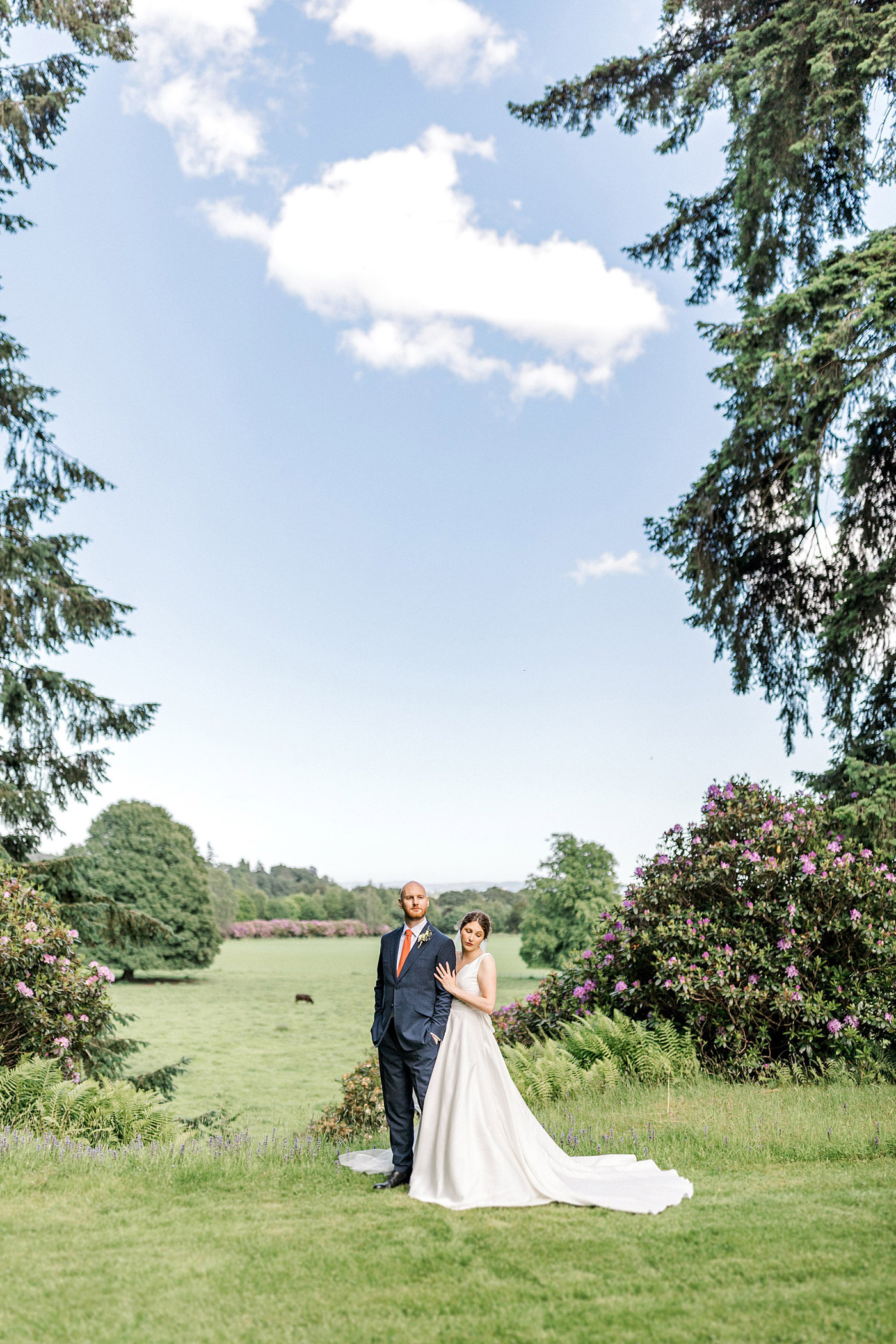 A bride in a white gown and a groom in a dark suit stand together on a lush green lawn, surrounded by tall trees and flowering shrubs, with an open field and blue sky in the background.