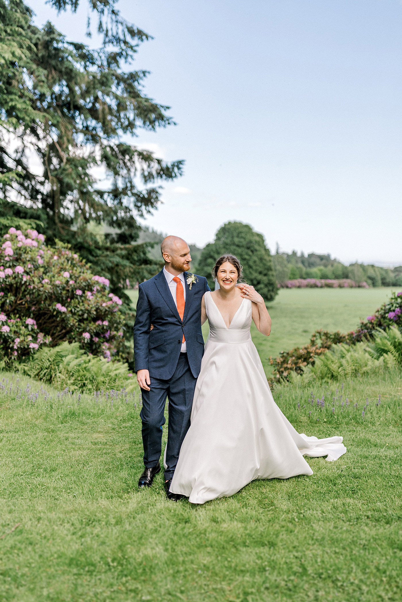 A bride in a white dress and a groom in a dark suit with an orange tie smile as they walk together on a grassy lawn, surrounded by blooming flowers and trees under a clear blue sky.