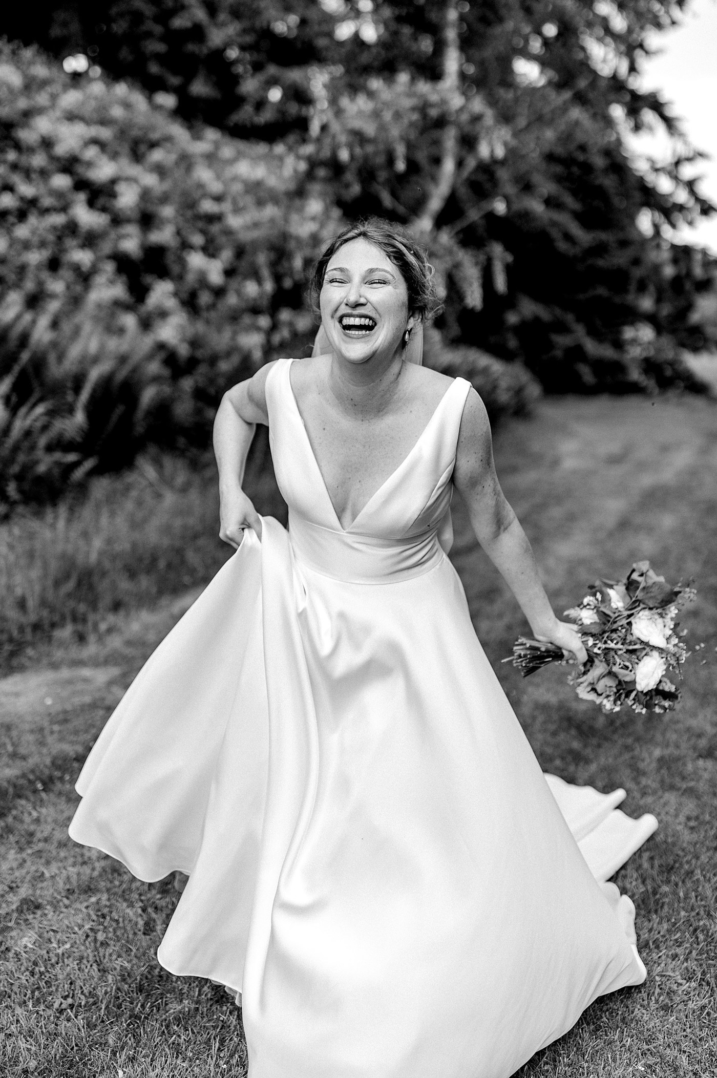 A joyful bride in a sleeveless wedding dress laughs whilst holding her bouquet and lifting her gown outdoors, surrounded by greenery.