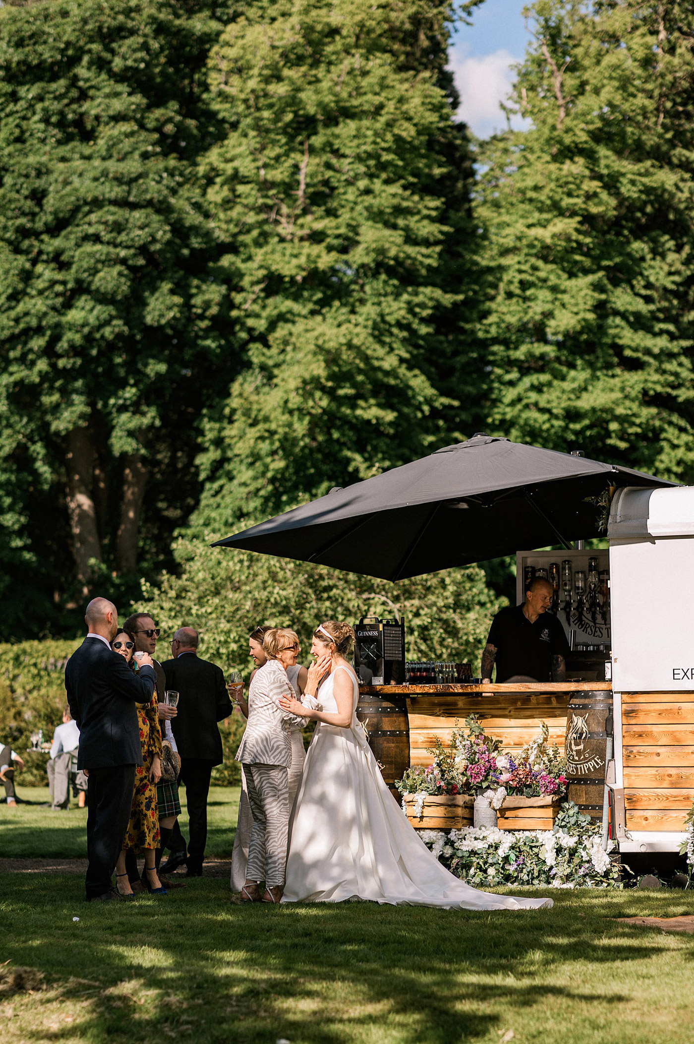 A bride in a white dress chats with guests near a rustic outdoor bar set up on a sunny lawn, surrounded by trees and decorated with flowers. Several people mingle and enjoy drinks under a large black parasol.