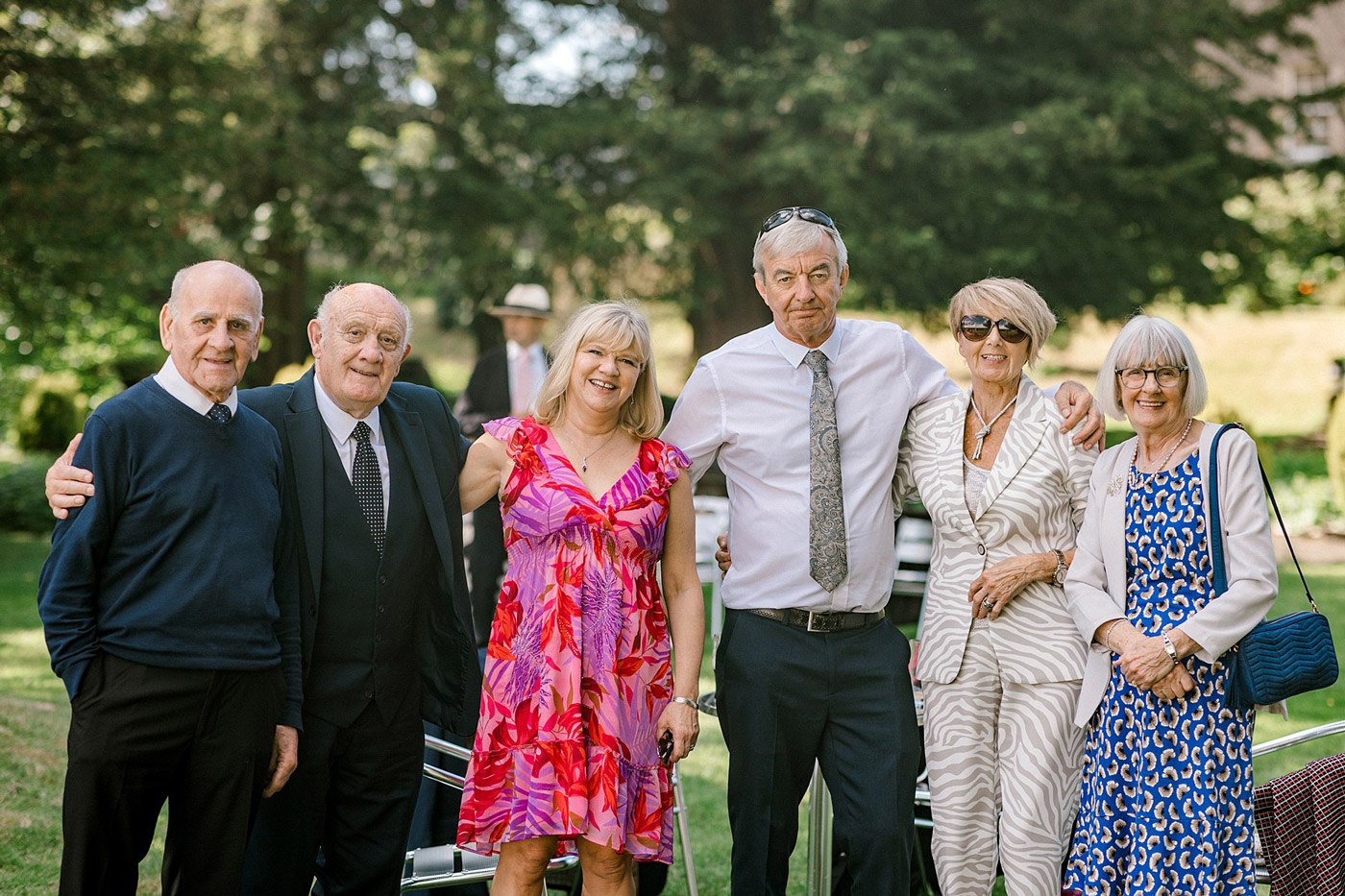 Six older adults stand together outdoors, smiling at the camera. The group is dressed in smart and colourful outfits, with greenery and trees in the background, suggesting a cheerful outdoor gathering or event.
